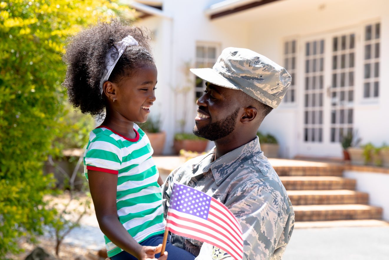 African American male solider wearing uniform holding his daughter with a USA flag in his arms, standing by the house on a sunny day, smiling and interacting.