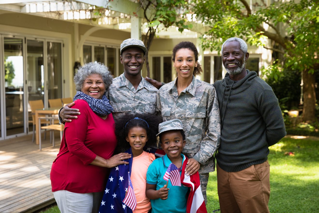 Portrait of a young adult African American male soldier and a young adult mixed race female soldier with thier diverse multi-generation family in the garden outside their home, embracing and smiling to camera, a US flag draped over the shoulders of the kids