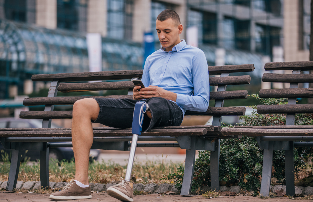 man with prosthetic leg sitting on bench outside looking at phone