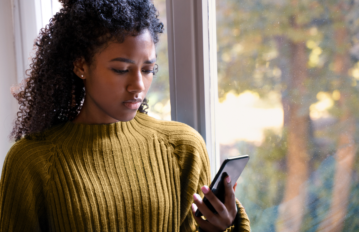 woman standing by window looking at phone with concerned look on face