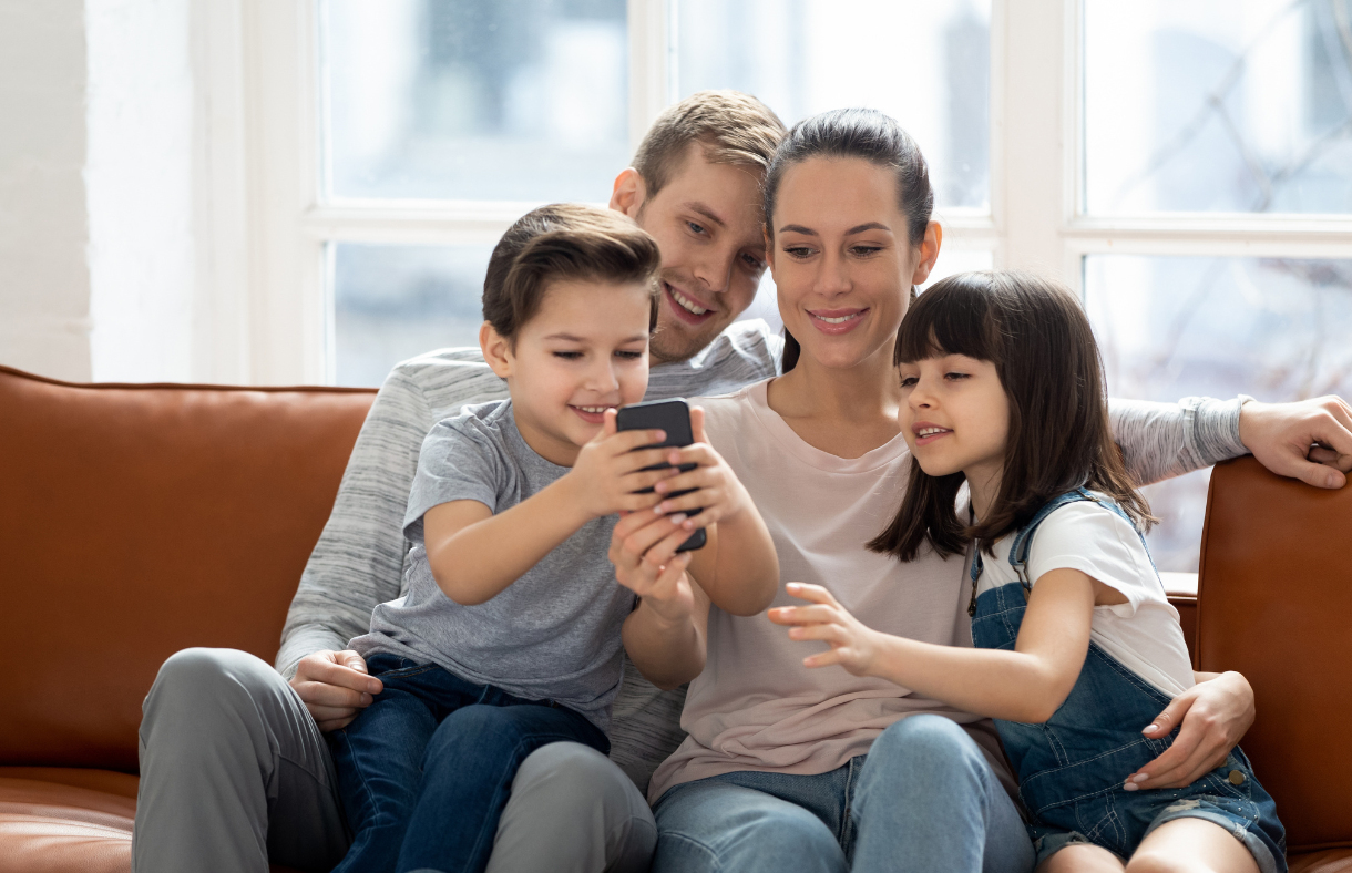 parents and two kids sitting on couch while all looking at phone together and smiling