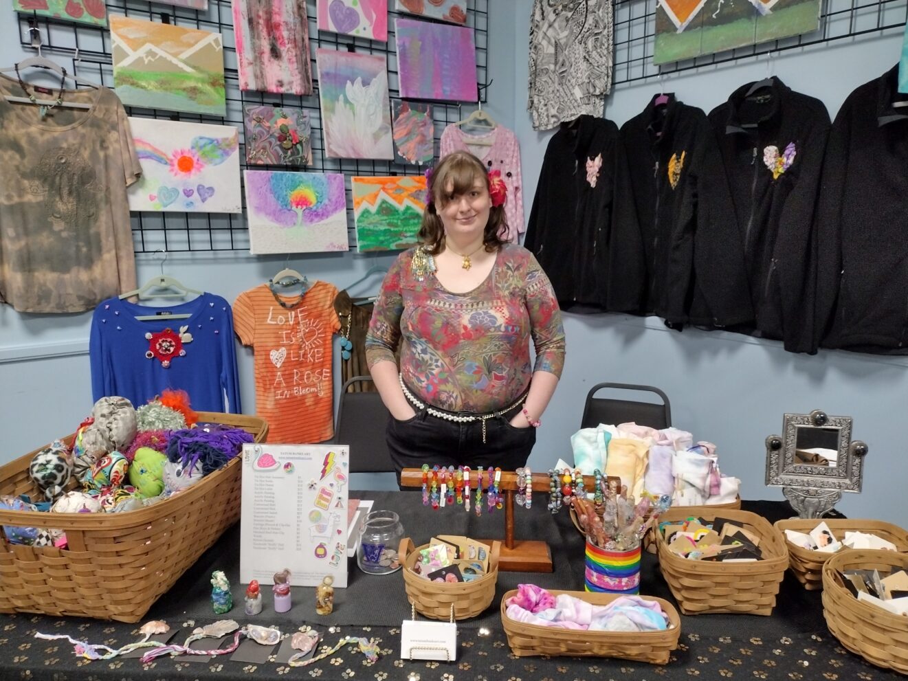 Tatum standing amid merchandise display: shirts, sweaters, and paintings hanging behind; jewelry, stuffed items, and other small crafts on table in front.