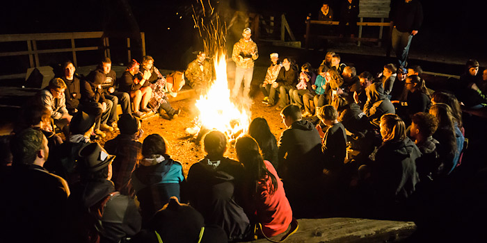 Campfire circle of about two dozen retreat participants, with leader standing and reading from a book.