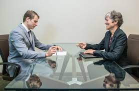 Two people facing each other across conference table, engaged in a practice job interview.