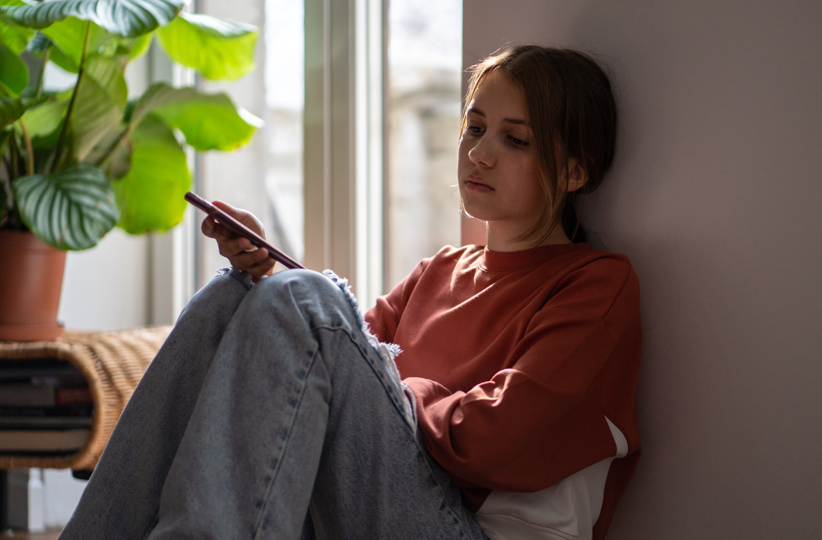 young adult girl with worried look on face sitting against wall next to window looking at phone