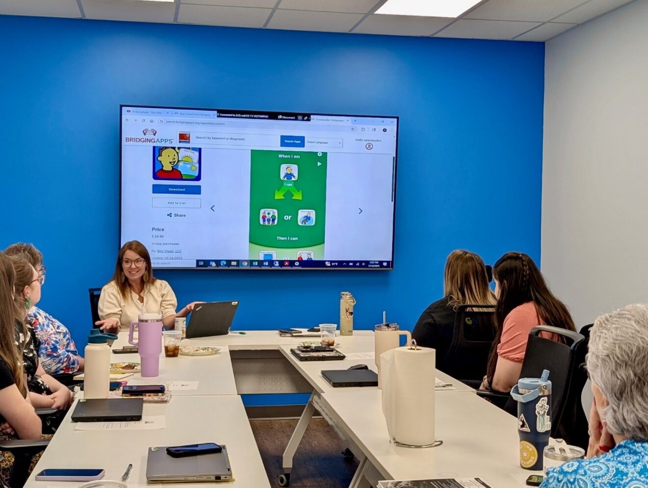 6 people seated around a rectangle of work tables, viewing a screen that shows the Choiceworks app navigating a smartphone-shaped green field.