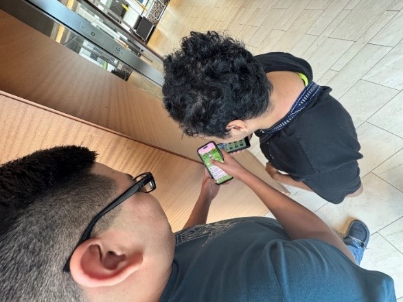 Photo of 2 students standing in corridor by brown-paneled corner wall. Both students are using smartphones. Both have short, dark hair and dark short-sleeved shirts; student on left is wearing black-framed glasses (visible only in extreme right profile; student faces are not showing).