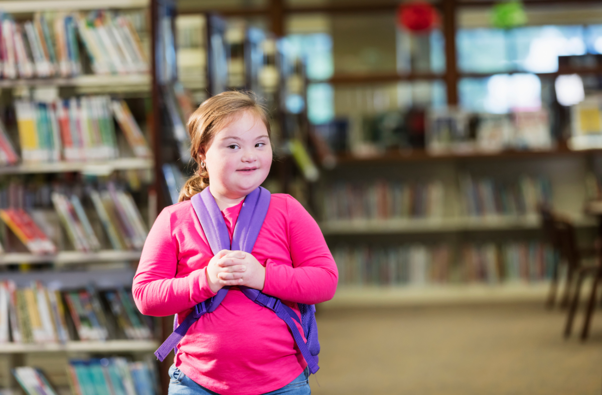 A child with Down syndrome wearing a pink jacket and blue backpack, smiling in a library.
