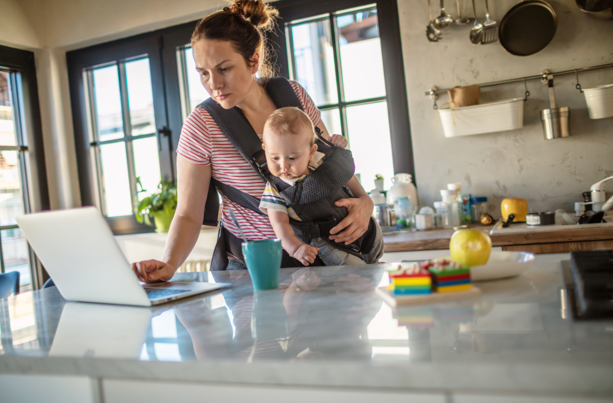 Woman wearing a baby carrier and holding a baby while working on a laptop in a kitchen. A coffee mug and toys are on the counter in front of her.