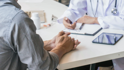 2 sets of male hands, one wearing medical coat, sitting at table with clipboard and ipad