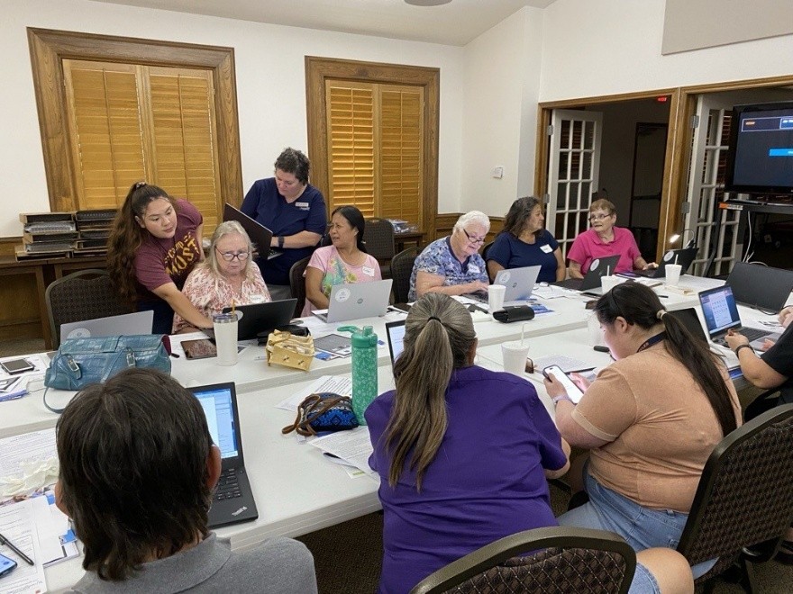 Group of people seated around a long, white table, using laptops. Two trainers, standing toward the left, are assisting participants.