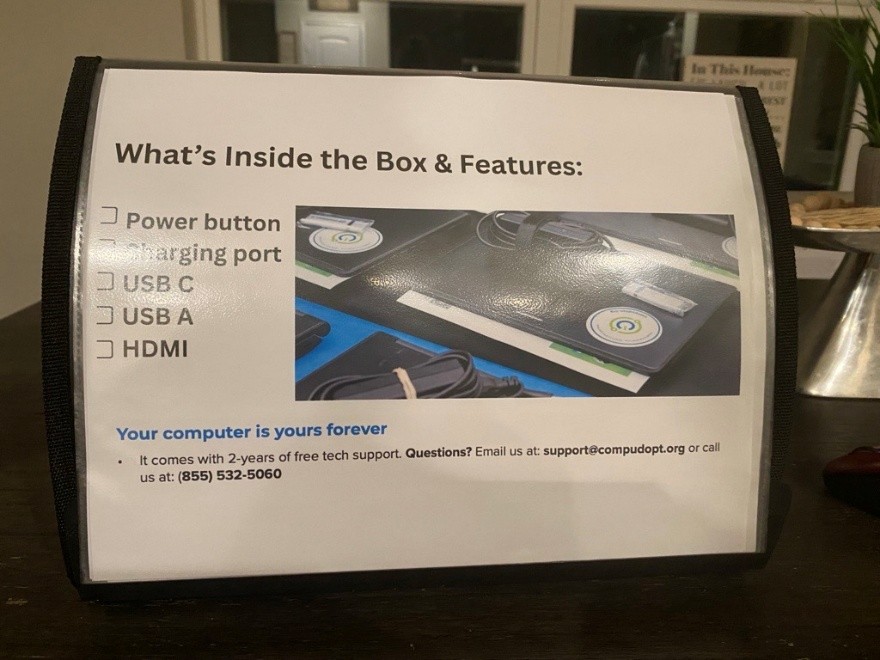 A table-tent-style page with a photo of technology gear on the right, surrounded by text: [Header] "What's Inside the Box & Features"; [Checklist] "Power button / Charging port / USB C / USB A / HDMI"; [Footer] "Your computer is yours forever. It comes with 2 years of free tech support. Questions? Email us at: support@compudopt.org or call us at: (855) 532-5060." 