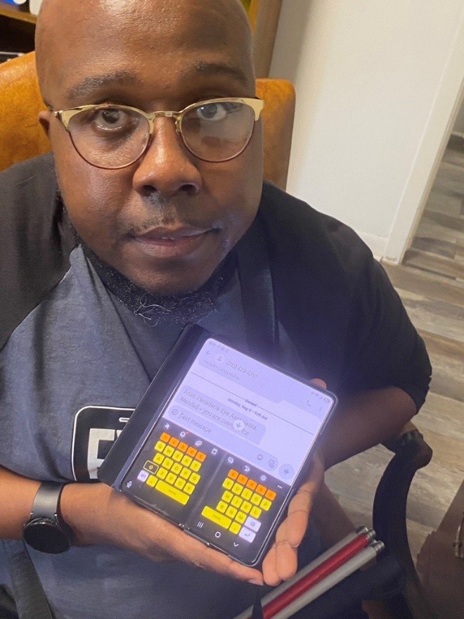 Close-up of young man in sports shirt and eyeglasses, holding up a small folding screen that displays a high-contrast keyboard in yellow, orange, and white.