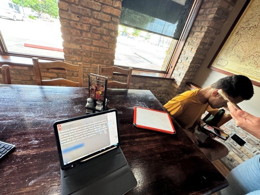 Indoor view of wooden table with one student seated to the right, and a hand holding a tablet visible in front of him. To the left, an open laptop displays a screen of live-captions instructions.