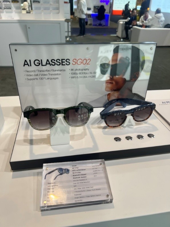 Close-up of expo table for "AI Glasses SG02." Displays two pairs of glasses with dark lenses and frames, an ad board standing behind, and a description card lying in front.