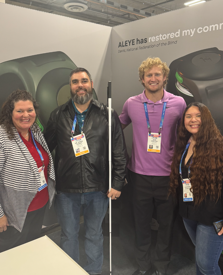 Four people, wearing CES 2026 lanyards, posing in front of a backdrop that features visual assistive technology. In order from left: (1) Female with long, wavy brown hair, wearing black-and-white striped blazer over red shirt. (2) Male with dark brown hair and beard, wearing black leather-type jacket, holding white cane in left hand. (3) Male with curly blond hair, wearing short-sleeved purple shirt. (4) Female with long dark hair, wearing dark shirt.