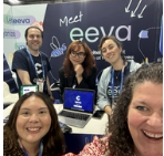 Head views of two people smiling in the foreground. In the background, a display backdrop reading "Meet eeva." Three people, visible from head to waist, standing behind a table in front of the backdrop. Laptop open on table, facing the camera.