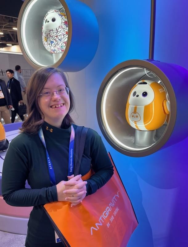 Sarah wearing a blue lanyard and holding an orange tote bag labeled "ANTIGRAVITY," standing next to a blue wall that displays two small egg-shaped robots in cubbyholes.