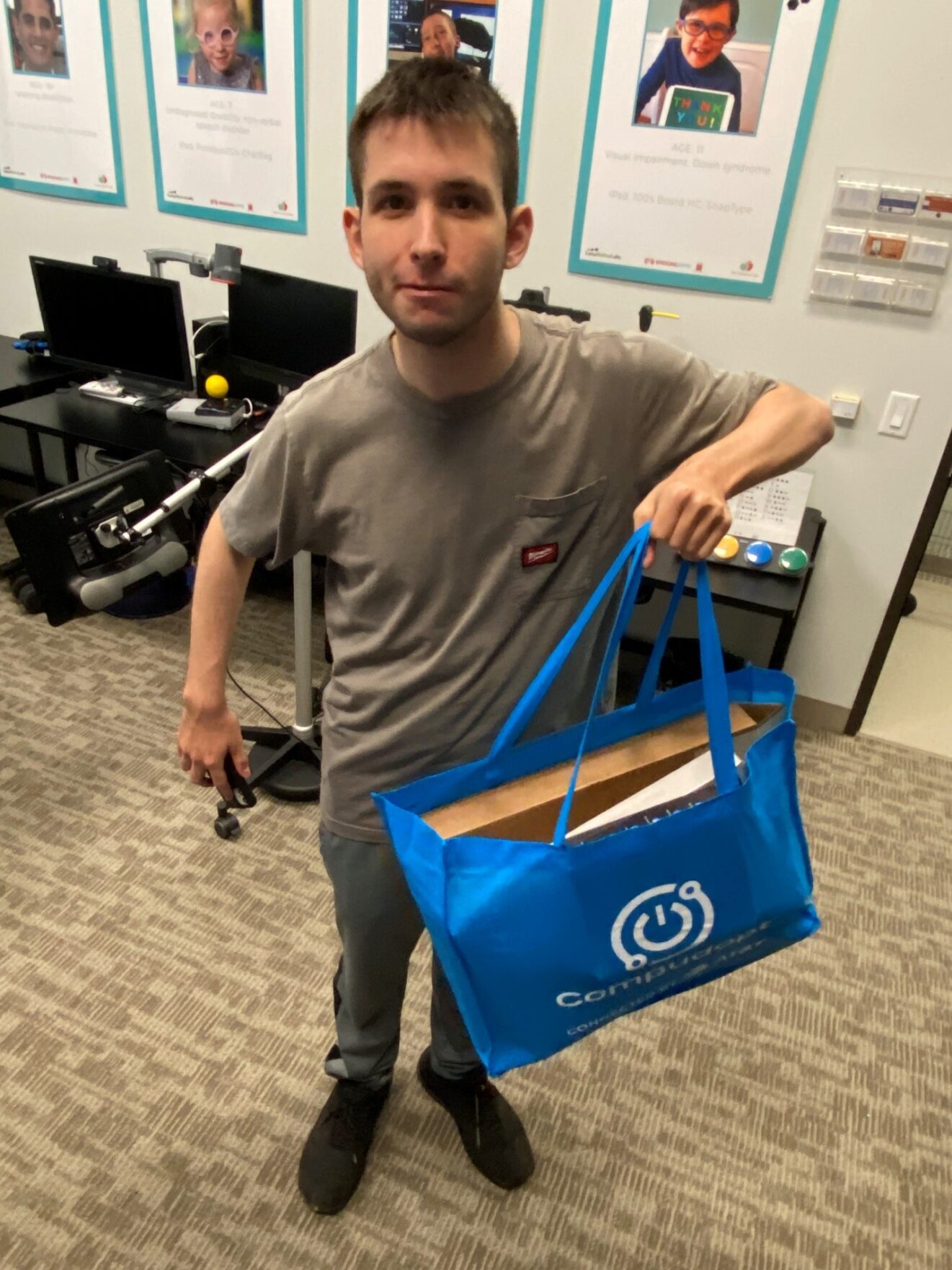 Full-length picture of Cameron Asaf standing in BridgingApps' assistive-technology lab. He has short, straight dark hair and is wearing a short-sleeved brown shirt with pants of matching color. He's holding a blue tote bag, with a Compudopt logo, in his left hand. A brown computer box is visible inside the bag.