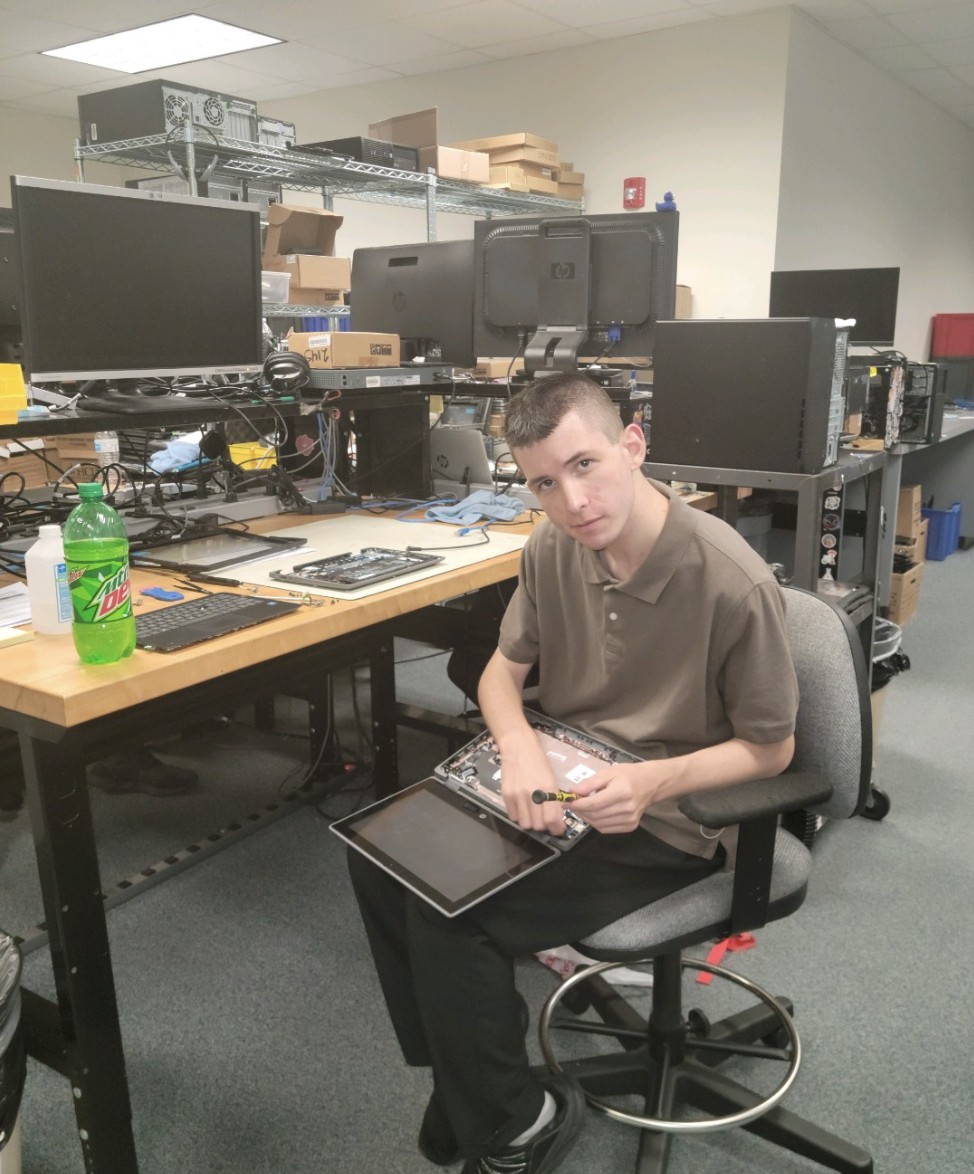 Cameron sitting in a room full of computers and equipment, holding a laptop with the keyboard removed for access to electronic parts.