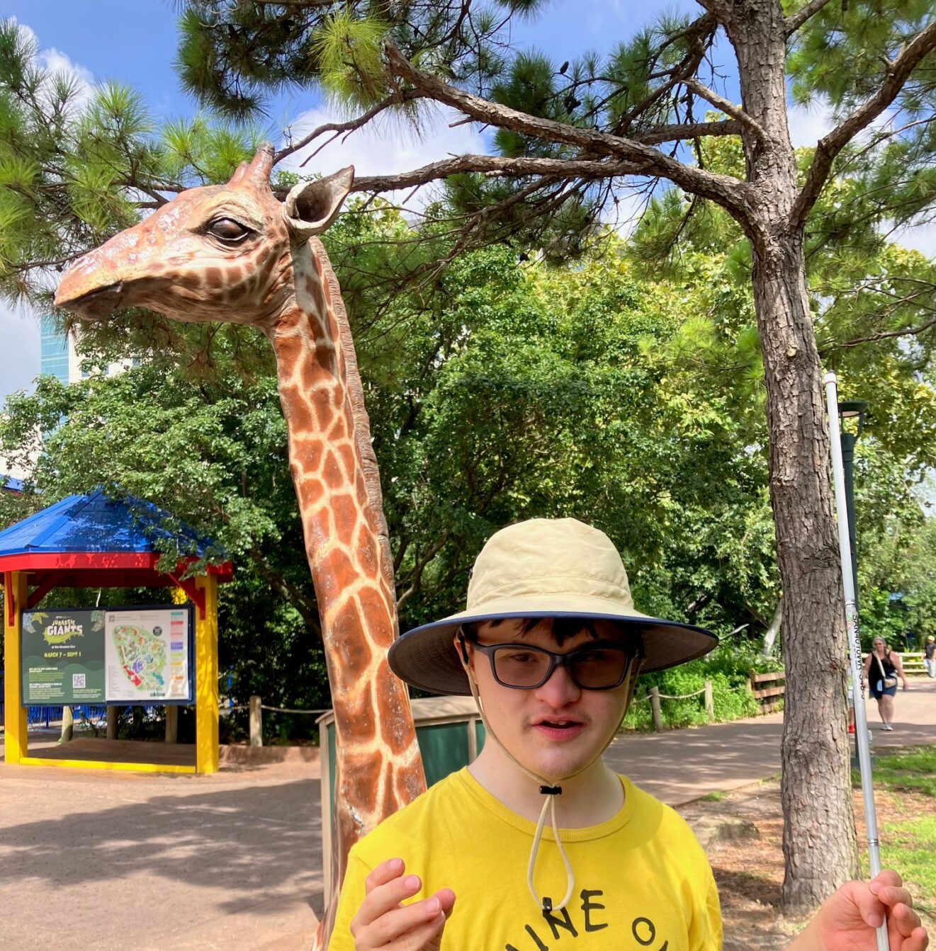 young adult guy wearing hat and yellow shirt standing in front of giraffe statue