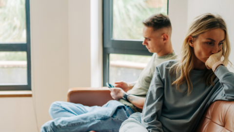 Two individuals sit on a couch in a room with large windows. One is looking at a phone, while the other appears deep in thought, with a hand over their mouth. The setting is calm and softly lit.