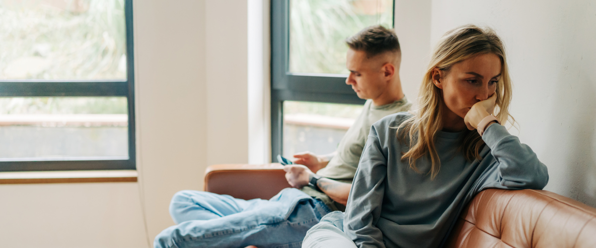Two individuals sit on a couch in a room with large windows. One is looking at a phone, while the other appears deep in thought, with a hand over their mouth. The setting is calm and softly lit.