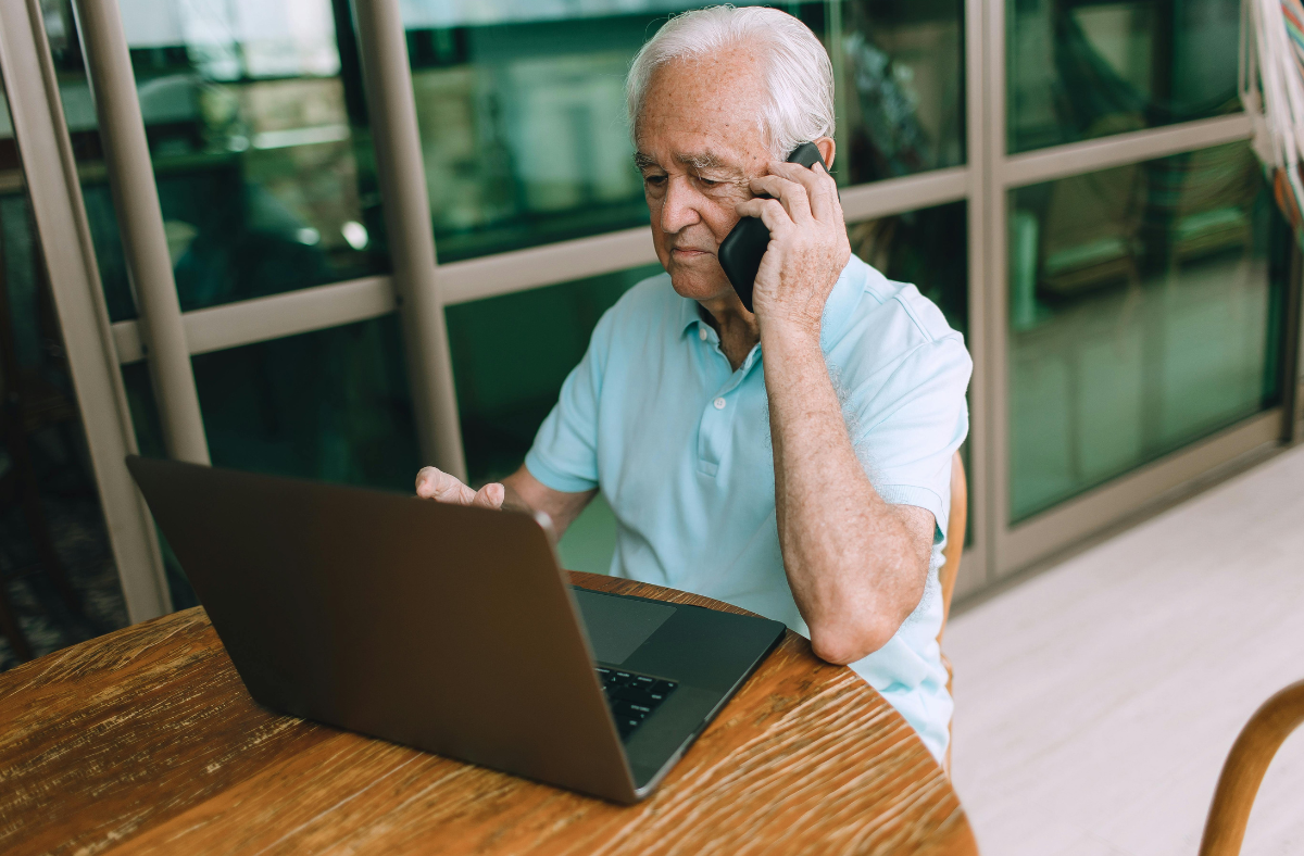 An individual with gray hair is sitting at a wooden table, using a laptop and holding a phone to their ear. They are in a room with large windows.