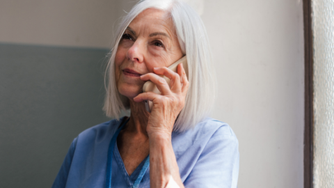 An adult with white hair is holding a phone to their ear, looking thoughtful, in a softly lit room with textured glass windows. They are wearing a blue top.