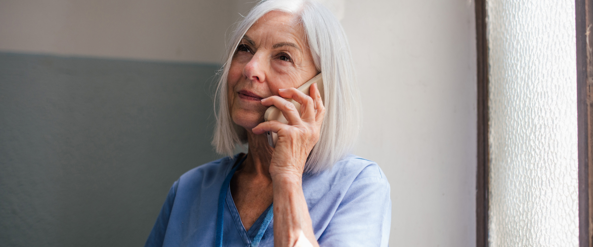 An adult with white hair is holding a phone to their ear, looking thoughtful, in a softly lit room with textured glass windows. They are wearing a blue top.