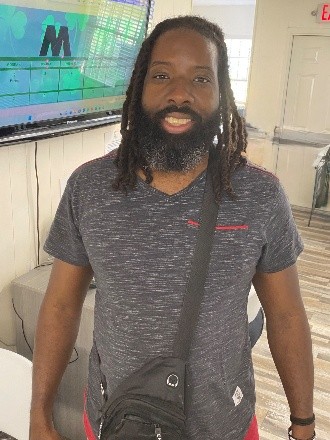 Head-to-hips front view of Hodari Patton, a young Black man with dreadlocks and a beard and moustache, wearing a short-sleeved gray shirt.
