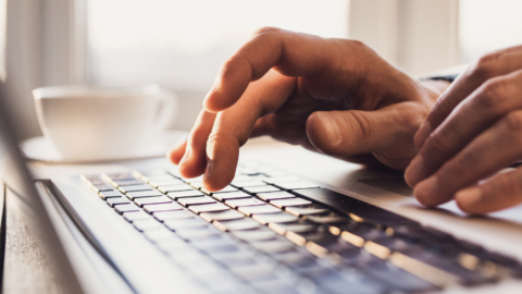 Hands typing on a laptop keyboard with a blurred cup in the background.