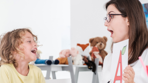 A person is holding a printed sheet with a large letter "A" and cherries, engaging with a young individual who is smiling. In the background, stuffed animals and art supplies are visible on shelves.