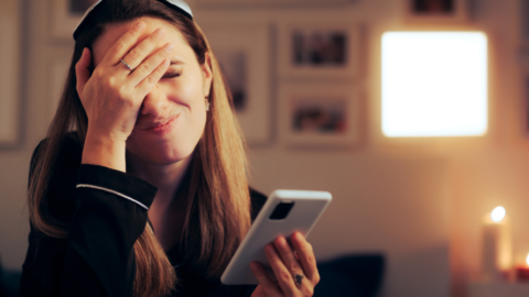 female looking stressed with one hand to her forehead and other holding phone in bedroom