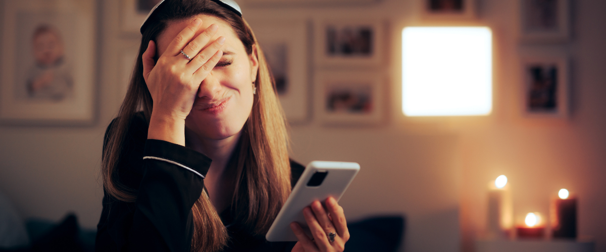 female looking stressed with one hand to her forehead and other holding phone in bedroom