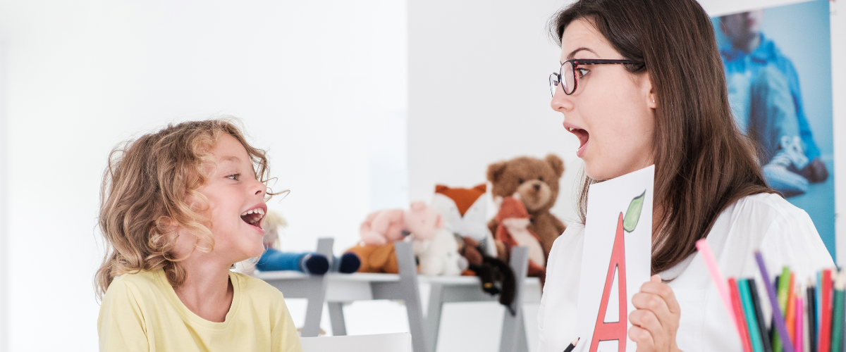 A person is holding a printed sheet with a large letter "A" and cherries, engaging with a young individual who is smiling. In the background, stuffed animals and art supplies are visible on shelves.