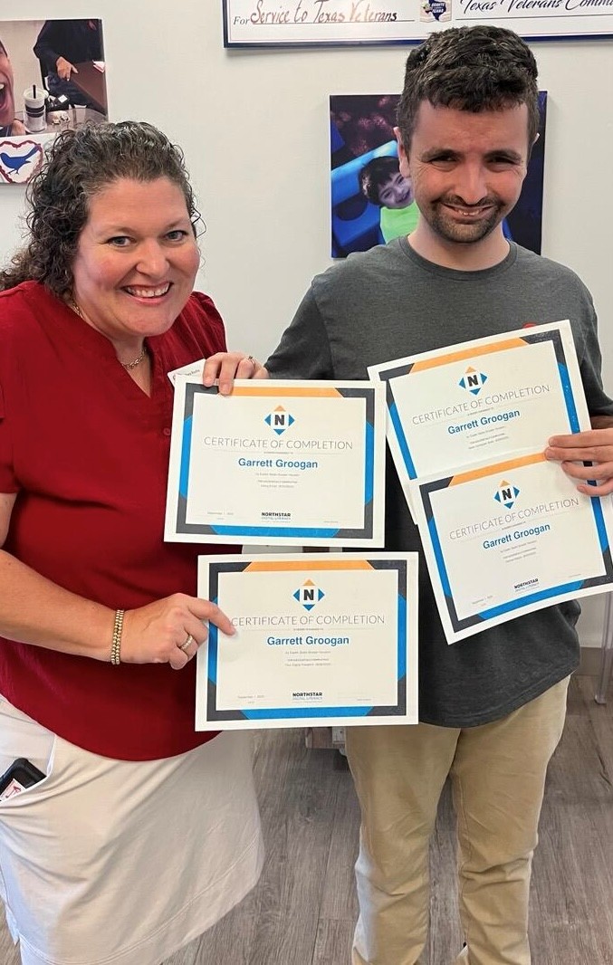 Close-up view. Tara Rocha (long curly hair, red shirt, white slacks) stands on left. Garrett (tall and slim, straight dark hair, small beard, gray shirt, khaki pants) stands on right. Each is smiling and holding up two "Certificates of Completion" with the Northstar logo on the top and Garrett's name in the center. 