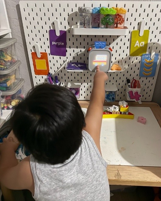 Child with short dark hair, back to camera, sitting at a work desk. Pointing to Yoto player (gray box showing digital image of van) on a small shelf to the right.