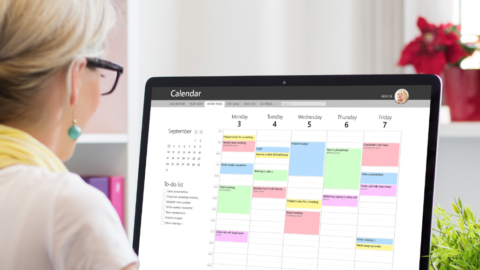 older woman seen from behind sitting at desk looking at computer screen showing digital calendar