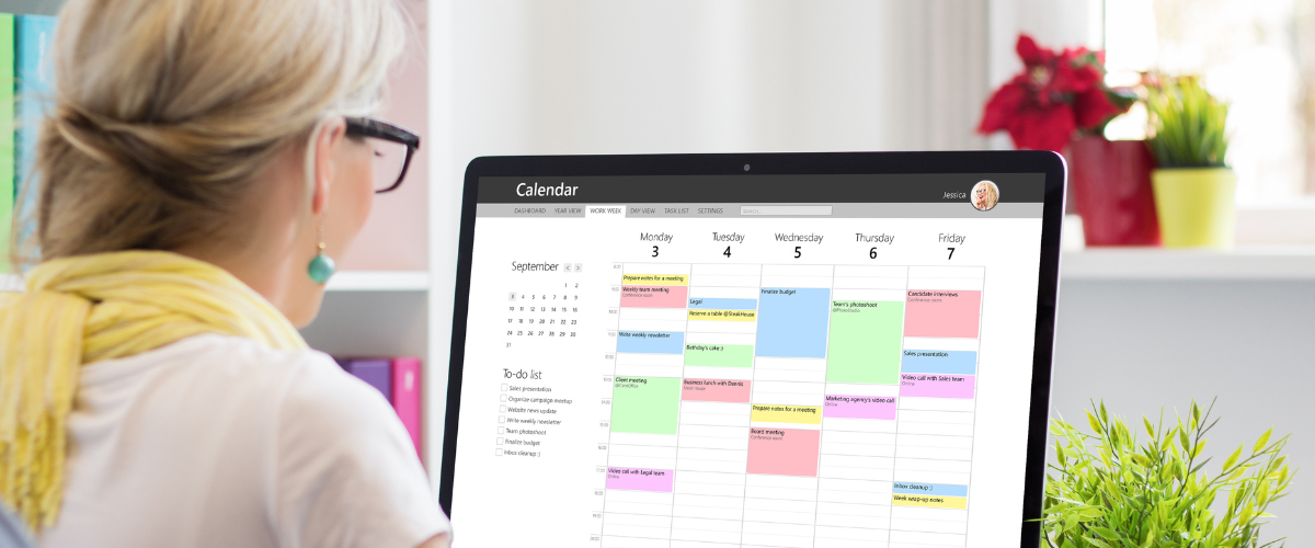 older woman seen from behind sitting at desk looking at computer screen showing digital calendar
