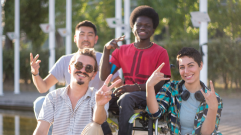 4 friends outside smiling and posing for camera, middle friend in sitting in wheelchair