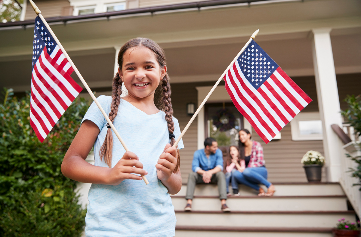 girl holding two american flags outside with family behind her sitting on porch