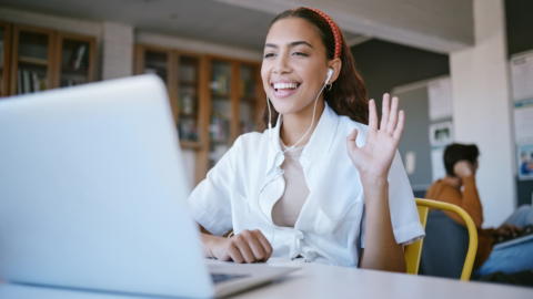 teen girl sitting in library with laptop smiling and holding up one hand