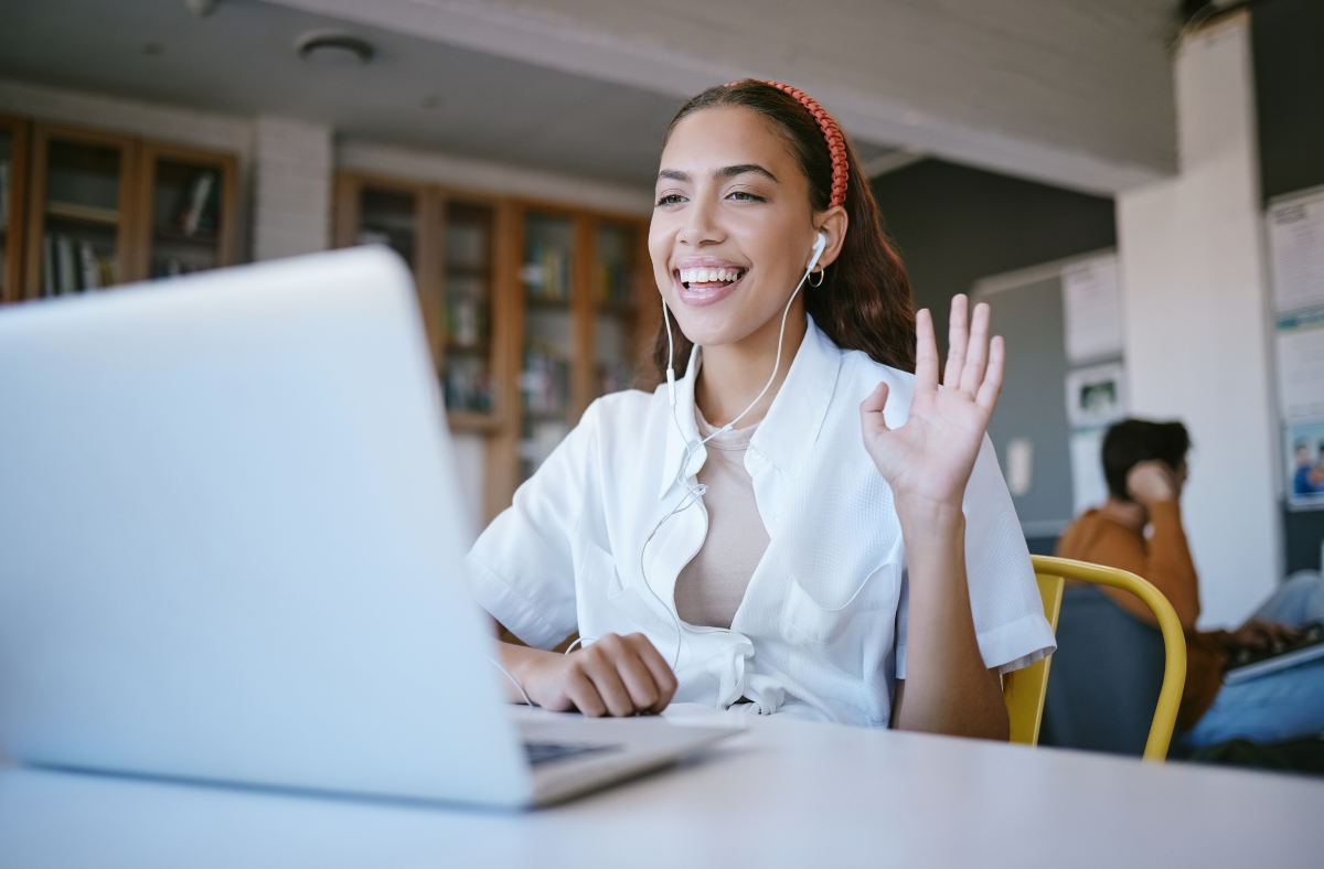 teen girl sitting in library with laptop smiling and holding up one hand