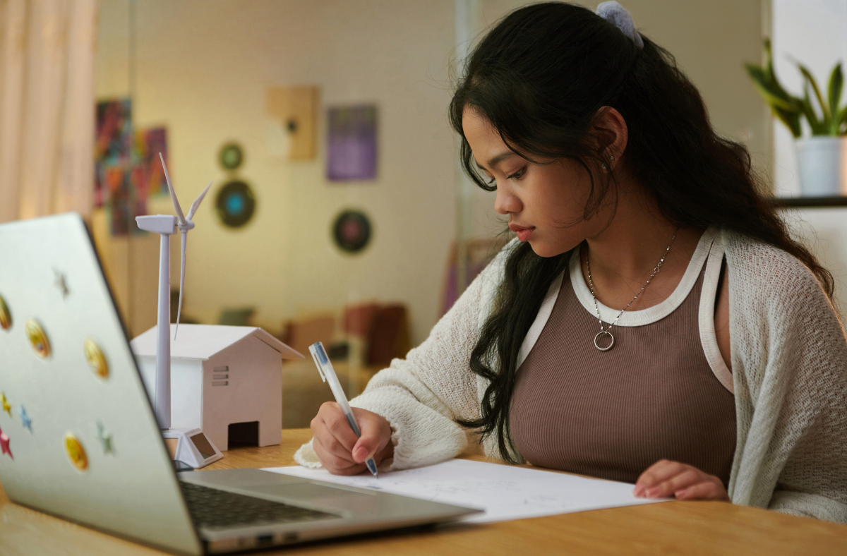 teen girl in room with open laptop writing on paper