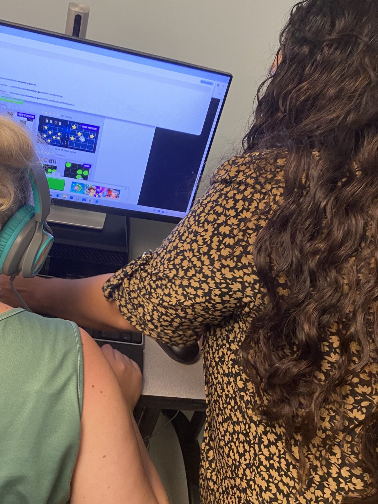 Ale, standing at right, helping client seated at left. Both are facing away from camera. Computer screen in front of client shows images against light blue background.
