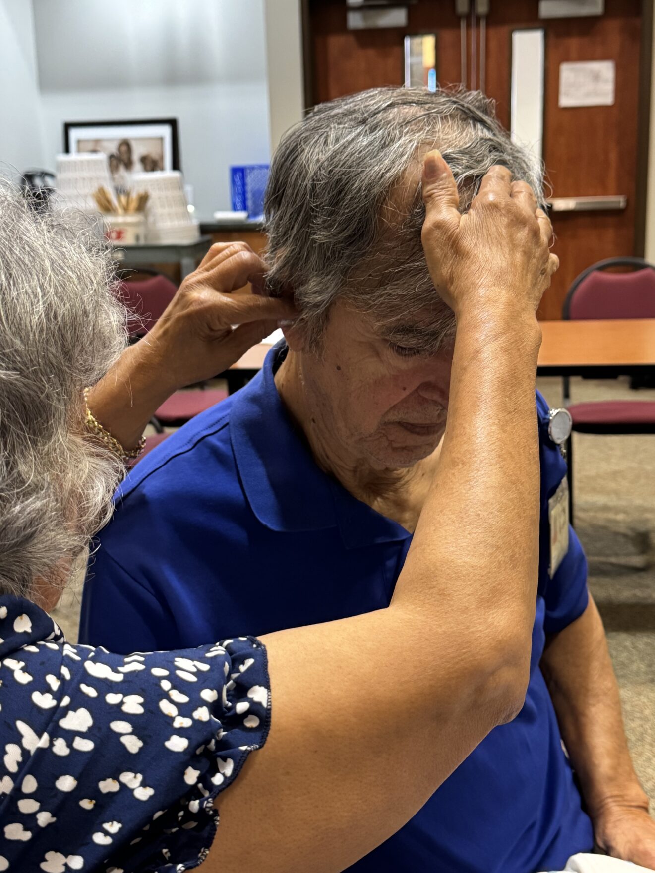 Person in navy-and-white blouse, back to camera, fitting hearing aid into right ear of man in blue shirt.