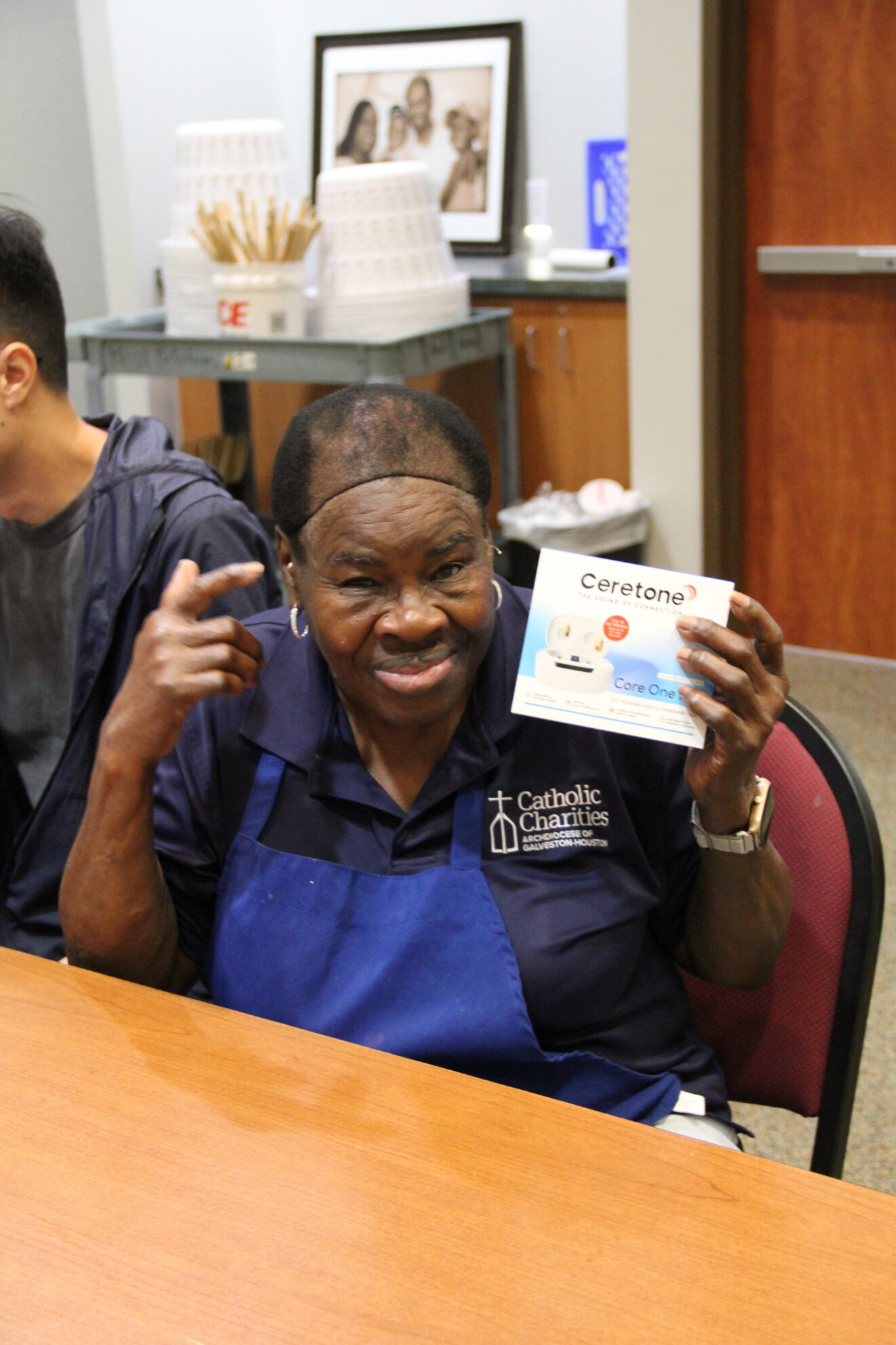 Woman wearing Catholic Charities jacket, black hairnet, and hoop earrings is holding up Ceretone box with her left hand and pointing to her right ear.