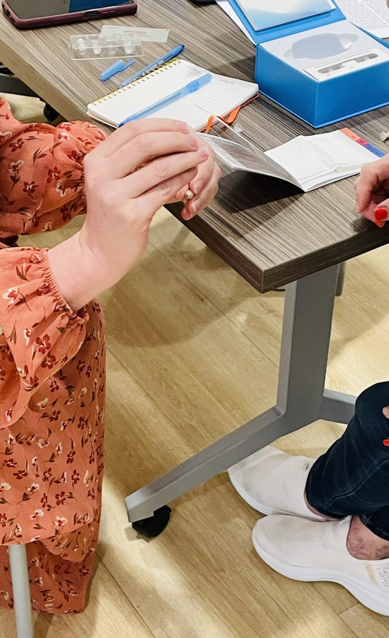Corner of rectangular table, on which are a blue box, two booklets, and other Ceretone-related items. Two people, partly visible, are seated facing each other (hands of the person on the left are showing, as are fingers and legs of the person on the right),