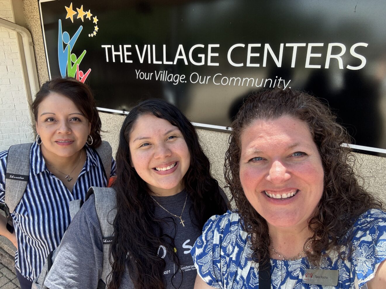 Silvia, Ale, and Tara lined up (left to right) in front of THE VILLAGE CENTERS banner. Banner has black background and shows Centers logo and slogan ("Your Village, Our Community").